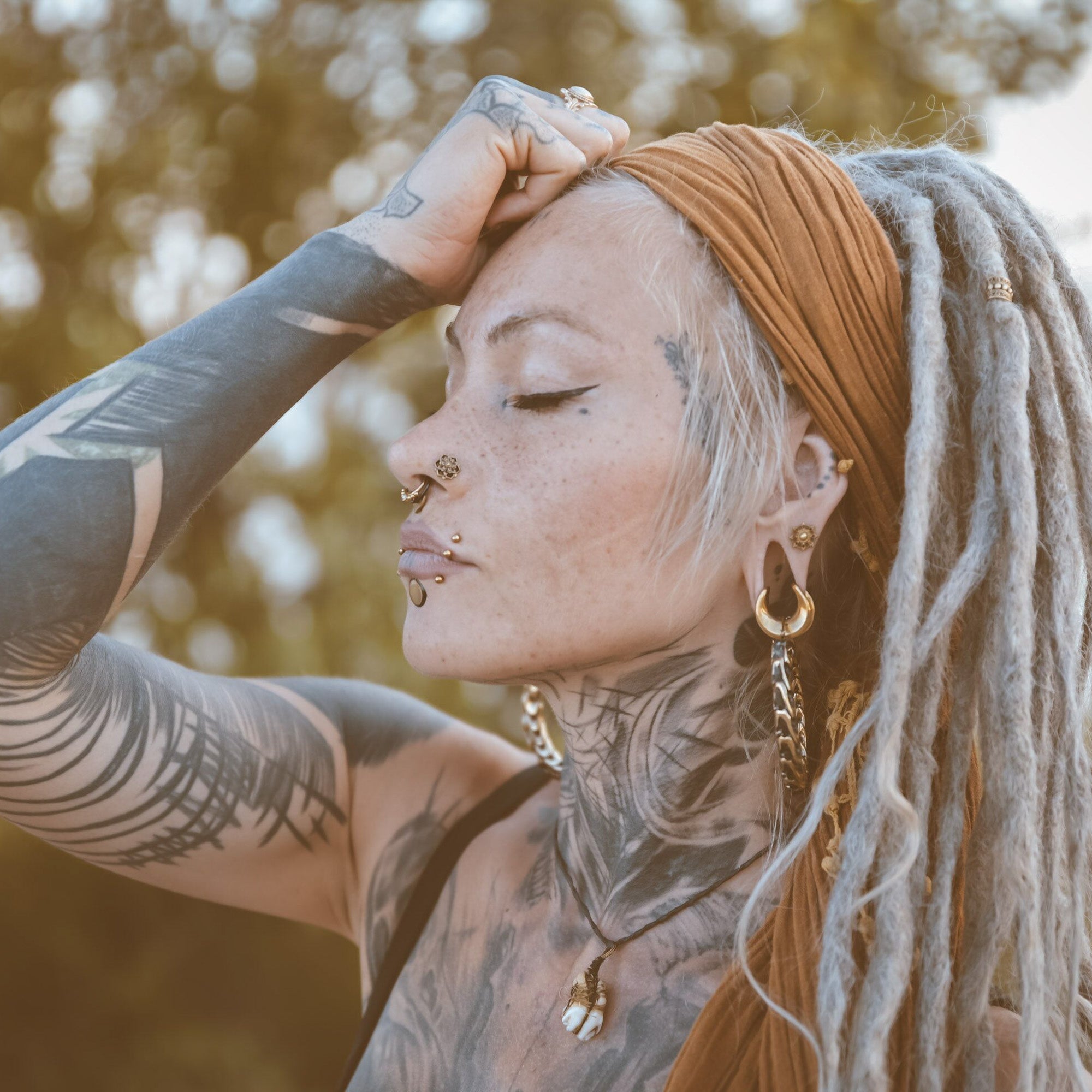 A person with long, grey dreadlocks and extensive tattoos on their face, neck, and arm is pictured outdoors. They wear a brown headwrap and post-apocalyptic body jewelry including multiple piercings and QUETZALCOATL Oversized Snake Spine Hoop Earrings in Gold (14 gauge) by Kaya Movement. Their eyes are closed, and one hand rests on their forehead, set against a backdrop of blurred foliage.